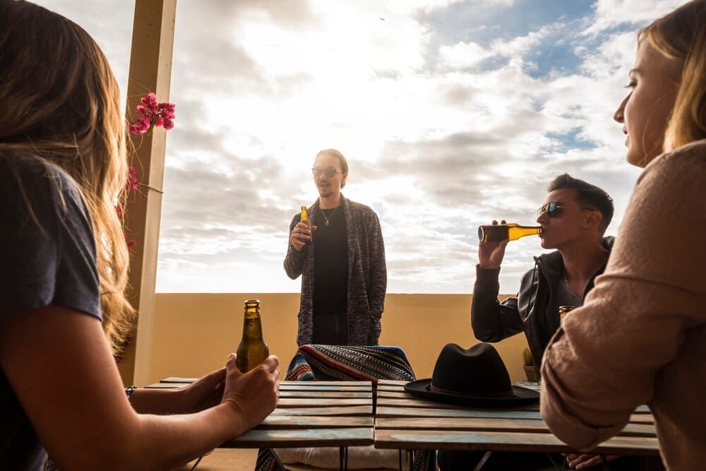 A group of young, white people socialize on a rooftop terrace, talking and drinking beer.