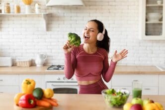 A smiling woman in workout clothes and earphones sings into a piece of broccoli in a kitchen.