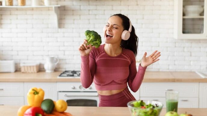 A smiling woman in workout clothes and earphones sings into a piece of broccoli in a kitchen.