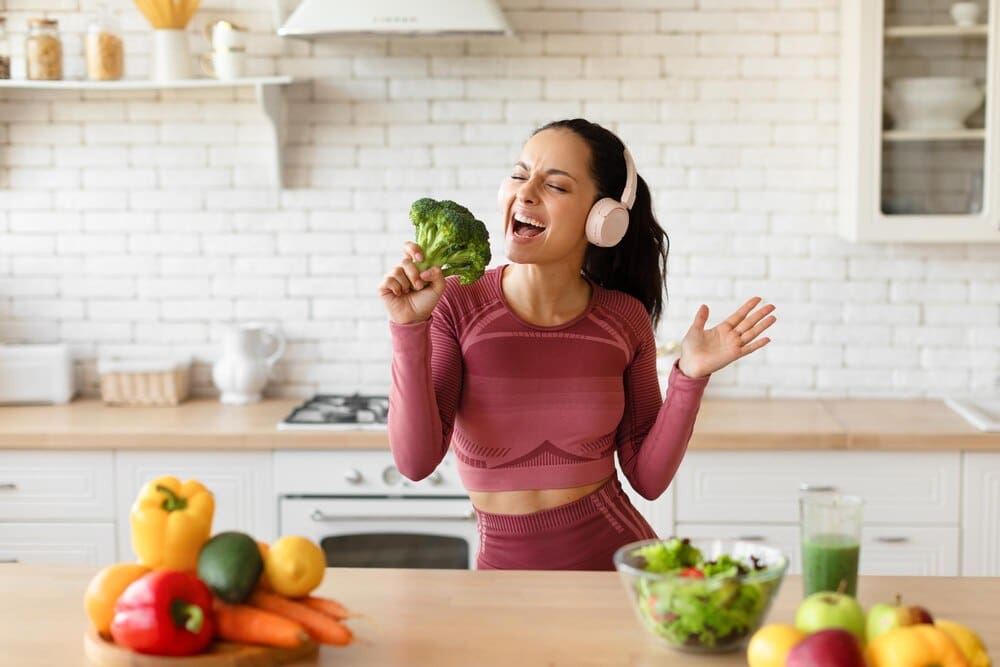A smiling woman in workout clothes and earphones sings into a piece of broccoli in a kitchen.