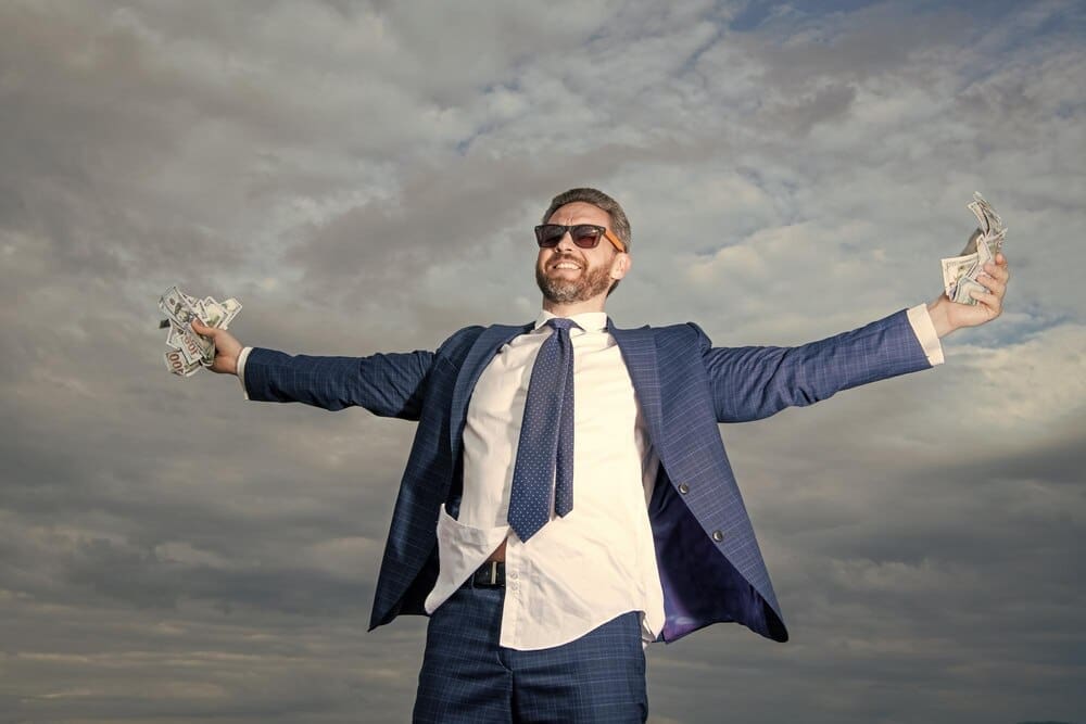 A smiling man in a suit holds money against a bright sky.