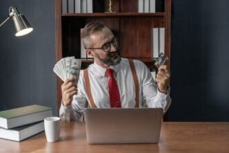 Happy senior businessman holding a stack of banknotes and a credit card, with a laptop on his desk.