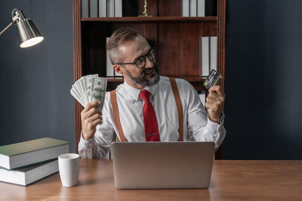 Happy senior businessman holding a stack of banknotes and a credit card, with a laptop on his desk.