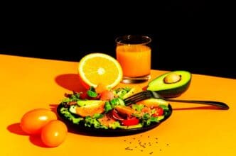 A still life photograph shows a healthy breakfast of salmon salad with avocado, sesame seeds, tomatoes, mixed herbs, and orange juice, set against an orange and black background.