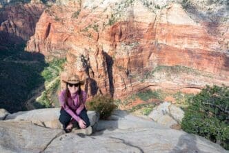 Woman crouches on a cliff, looking out over Zion Canyon from a high angle perspective.
