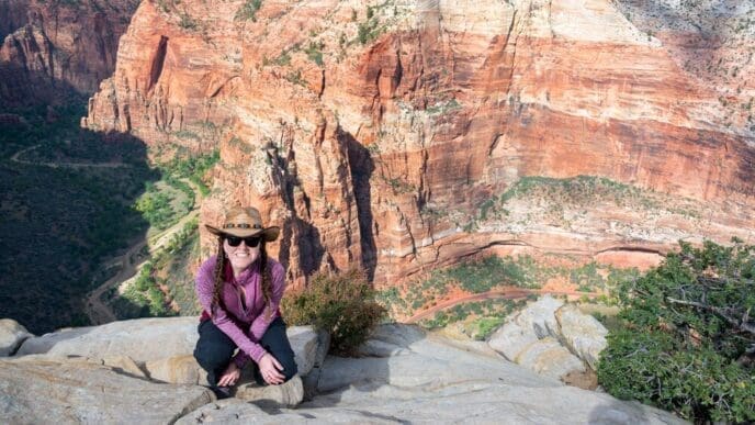 Woman crouches on a cliff, looking out over Zion Canyon from a high angle perspective.