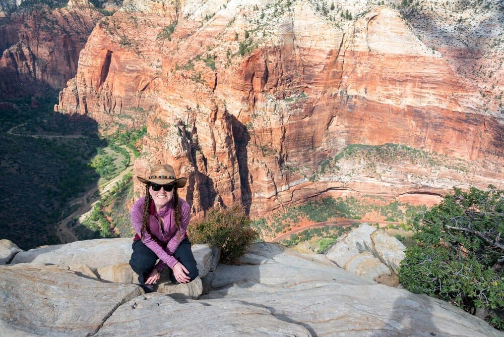 Woman crouches on a cliff, looking out over Zion Canyon from a high angle perspective.