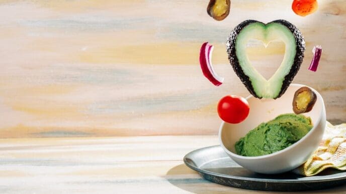 A bowl of homemade guacamole is surrounded by its ingredients on a vintage wooden table.