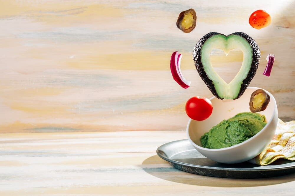 A bowl of homemade guacamole is surrounded by its ingredients on a vintage wooden table.
