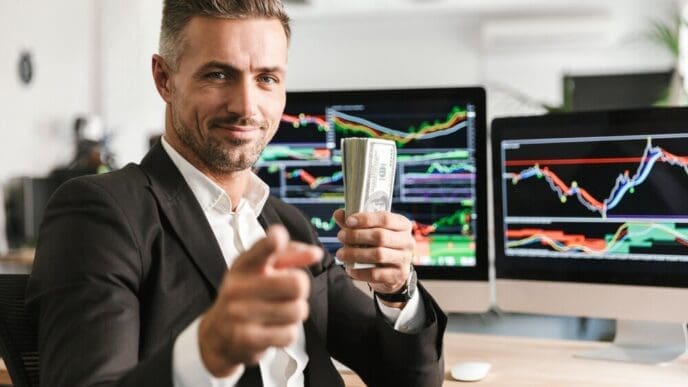 A well-dressed businessman in his 30s holds a stack of money while working on a computer in an office with financial charts.