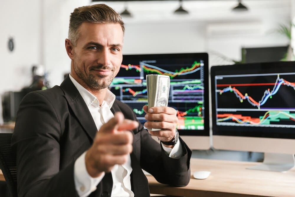 A well-dressed businessman in his 30s holds a stack of money while working on a computer in an office with financial charts.