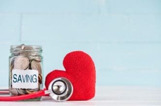 A glass jar filled with coins sits on a wooden surface, with a stethoscope and a small red heart resting nearby.