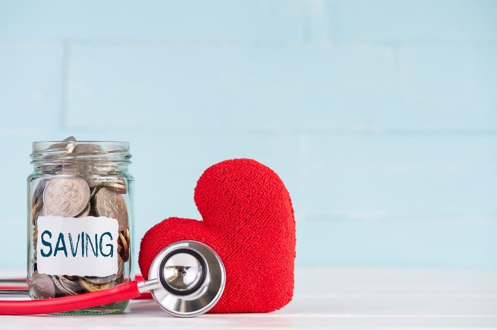 A glass jar filled with coins sits on a wooden surface, with a stethoscope and a small red heart resting nearby.