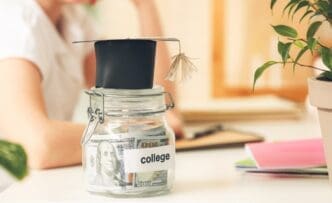 A glass jar filled with coins sits on a table next to a graduation cap, symbolizing savings for education.