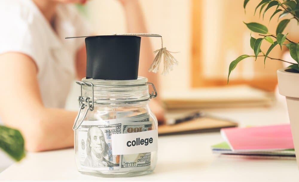 A glass jar filled with coins sits on a table next to a graduation cap, symbolizing savings for education.