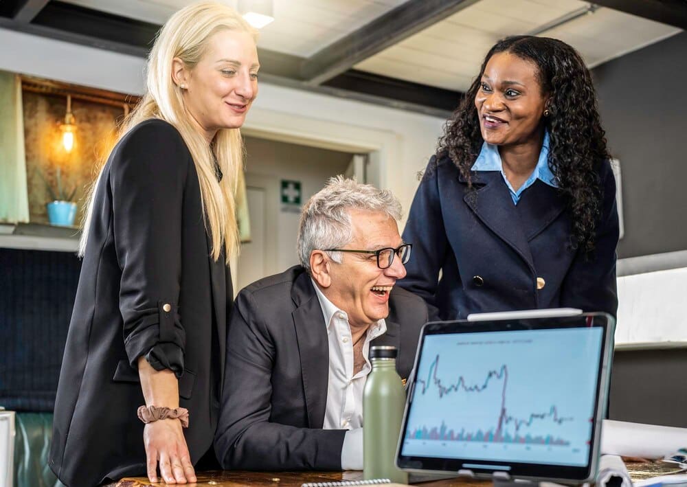 A diverse business team smiles while reviewing market trends on a computer during a meeting.