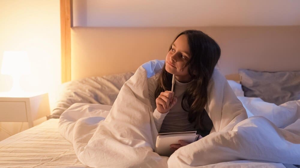 A small girl lies in bed, deep in thought, perhaps inspired by art or a hobby.