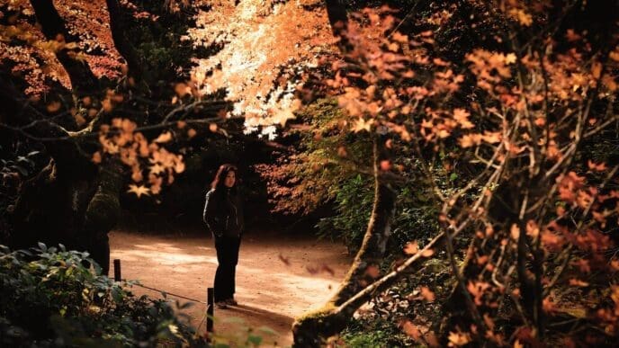 A woman stands among colorful autumn leaves in a traditional Japanese garden.