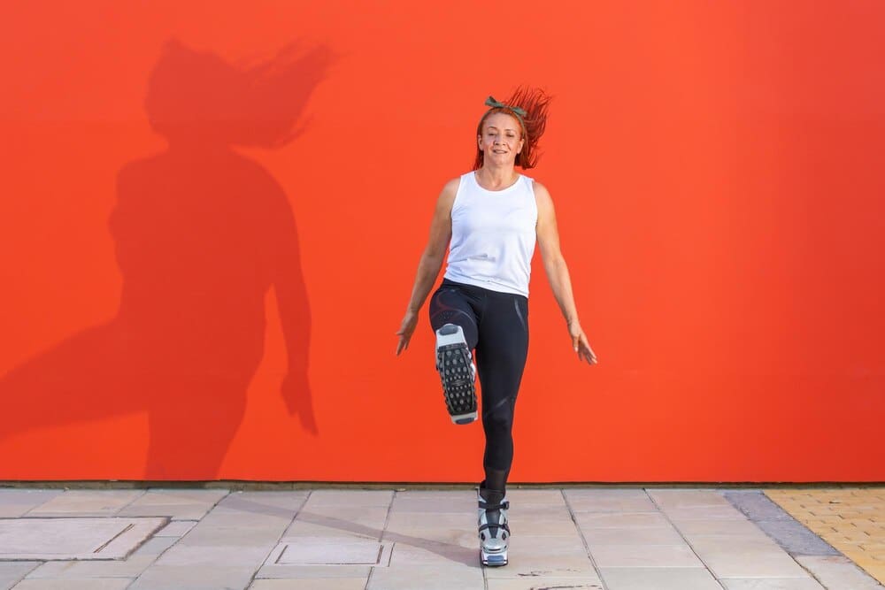 A Latina redhead woman in sportswear exercises with Kangoo Jumps boots on an orange background.