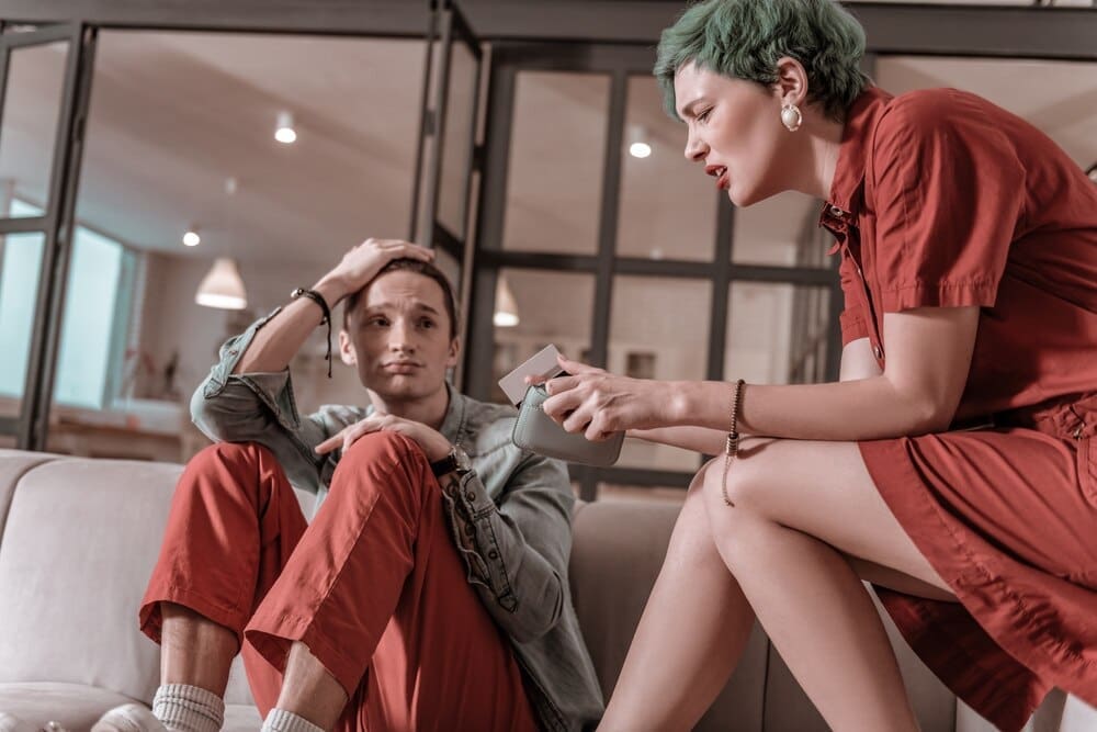 A green-haired woman looks surprised and disappointed while holding bank cards, likely indicating a lack of funds.