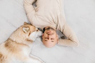 A man laughs while lying on the floor next to a fluffy husky dog.
