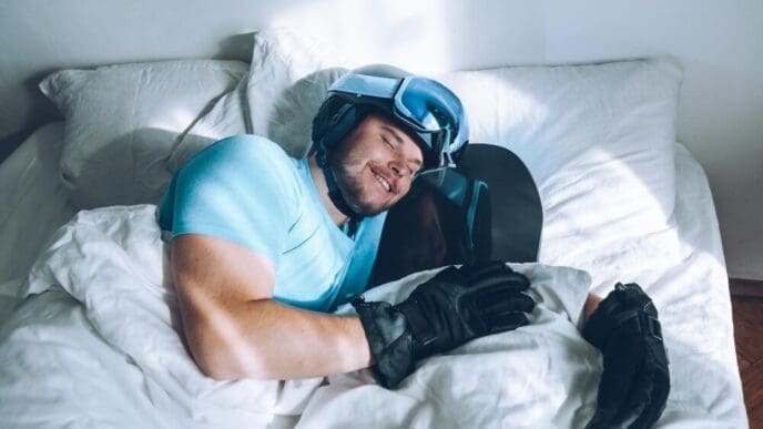 Man wearing a snowboard helmet and goggles lies in bed.