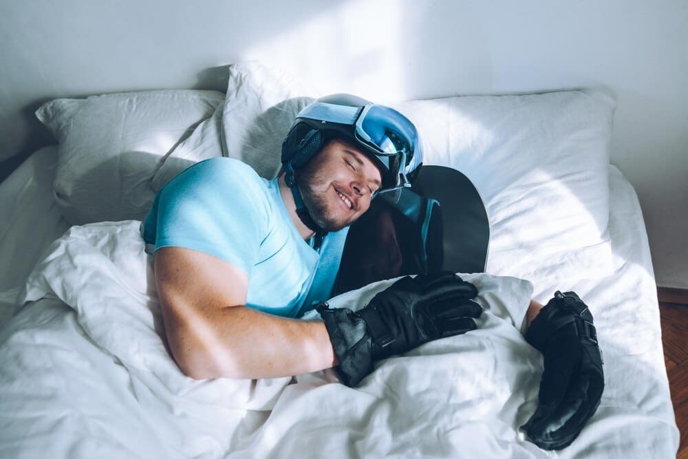 Man wearing a snowboard helmet and goggles lies in bed.