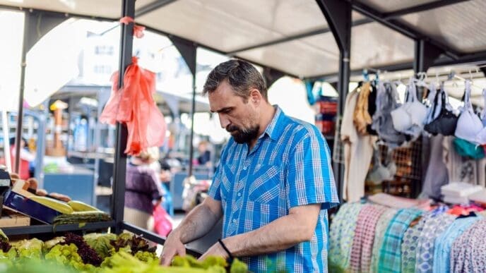 A man with a beard and plaid shirt selects vegetables at a market.