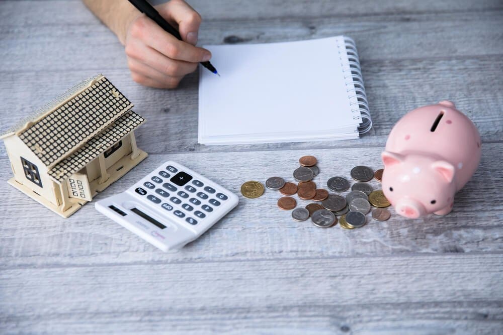 A man's hand holds a notepad with coins and a piggy bank in the background.