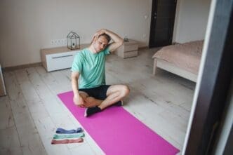 A man sits cross-legged on a fitness mat, stretching his neck in preparation for a workout.