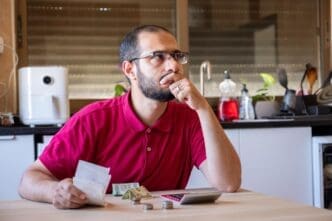 A man sits at a kitchen table, calculating expenses with a thoughtful expression.