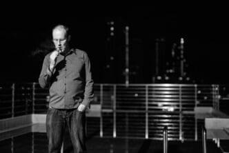 A man stands near a railing at night, likely overlooking a cityscape or landscape.
