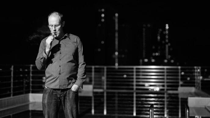 A man stands near a railing at night, likely overlooking a cityscape or landscape.