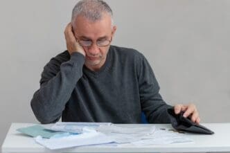 A mature man sits at home, looking stressed and confused, likely due to financial worries.