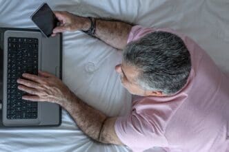 Mature man with a beard is working on a laptop while sitting in bed.