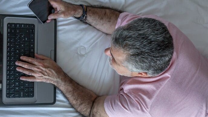 Mature man with a beard is working on a laptop while sitting in bed.
