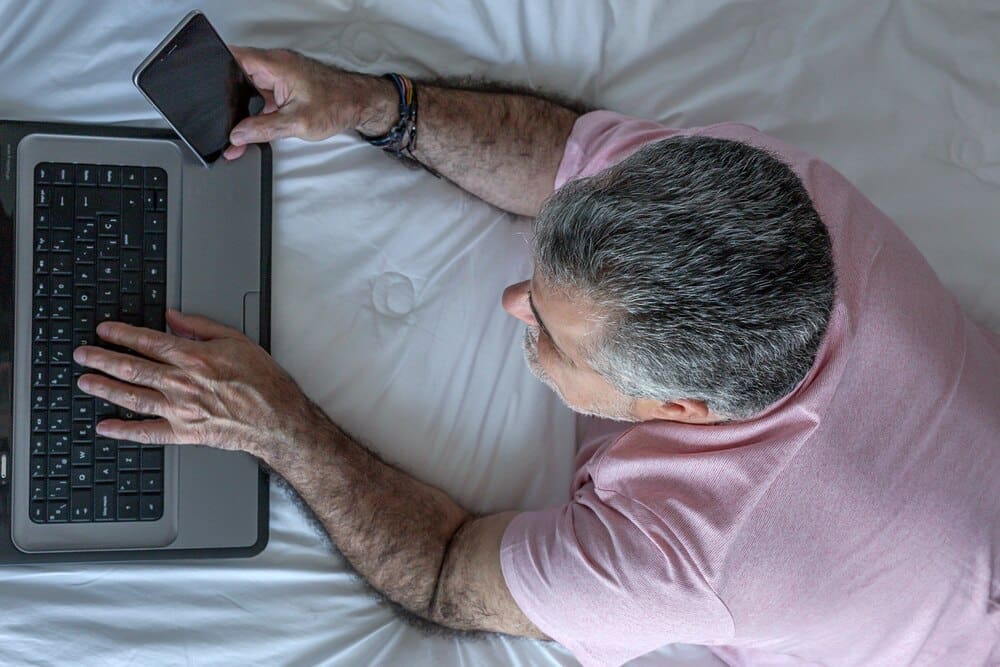 Mature man with a beard is working on a laptop while sitting in bed.