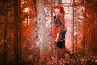 A mature model with red curly hair poses as a royal huntress, aiming a crossbow in a forest setting.