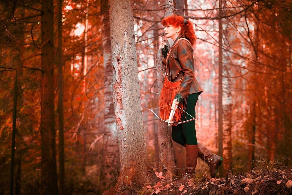 A mature model with red curly hair poses as a royal huntress, aiming a crossbow in a forest setting.