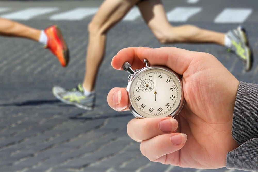 A close-up shows a mechanical stopwatch with a hand measuring speed, with a runner's legs blurred in the background.