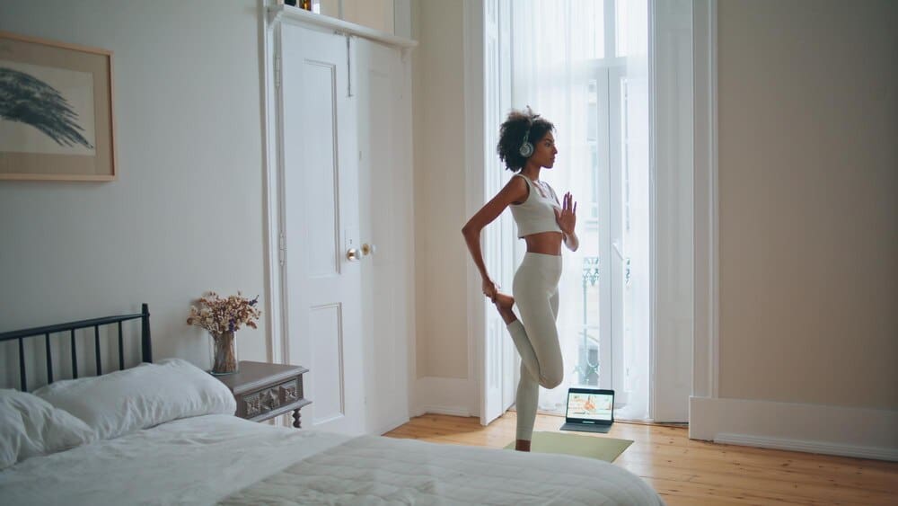 A dark-skinned woman in a bedroom meditates while performing a yoga asana.