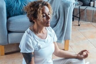 A middle-aged woman sits cross-legged on a carpet in her living room, eyes closed, meditating in a lotus position.
