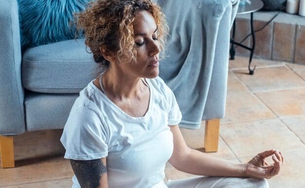 A middle-aged woman sits cross-legged on a carpet in her living room, eyes closed, meditating in a lotus position.
