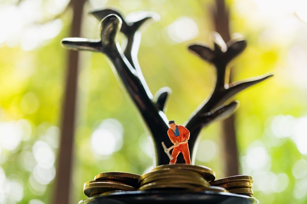 Miniature figures work on a stack of coins beneath a tree-like structure, symbolizing business concepts.