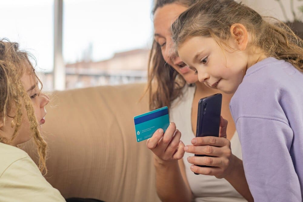 A mother with a bank card and smartphone demonstrates financial technology to her two young sons with dreadlocks.
