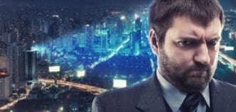 A somber businessman stands in front of a brightly lit cityscape at night.