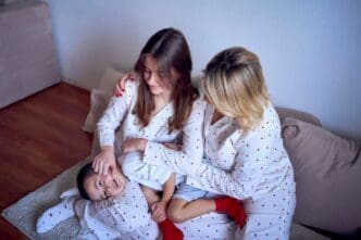 A mother and teenage girl in pajamas tickle a younger boy on the floor.