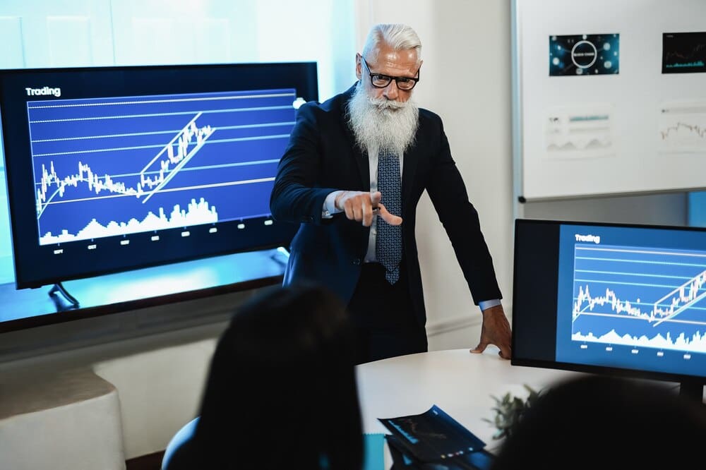 A diverse group of stock traders, with a focus on an older man, collaborate on market analysis in a busy office setting.