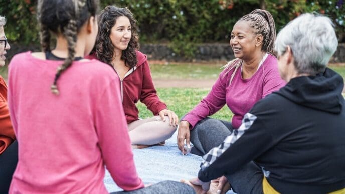 A smiling senior Black woman is the focal point of a multi-generational group of women enjoying themselves outdoors in a city park.