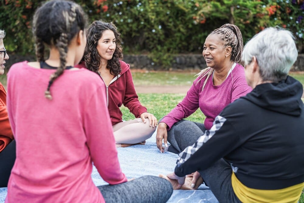 A smiling senior Black woman is the focal point of a multi-generational group of women enjoying themselves outdoors in a city park.